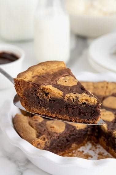 A slice of brookie pie being lifted from a white porcelain pie plate on a metal spatula.