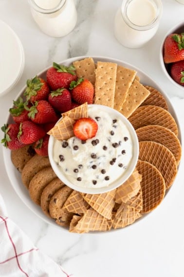 A white serving platter filled with cookies, graham crackers, strawberries and sugar cone wafers. In the center of the dish is a white bowl filled with chocolate chip cannoli dip.