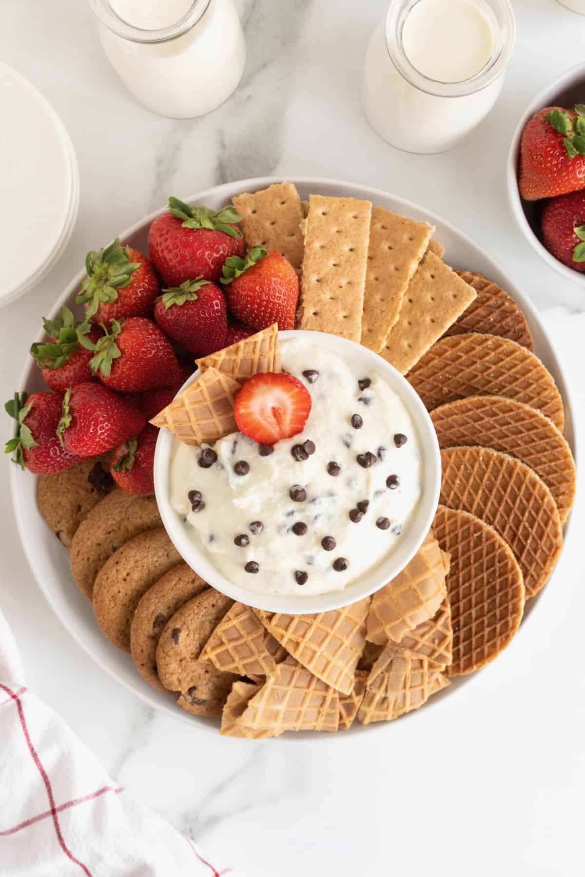 A white serving platter filled with cookies, graham crackers, strawberries and sugar cone wafers. In the center of the dish is a white bowl filled with chocolate chip cannoli dip.