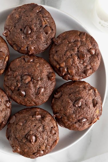 A white serving plate with seven chocolate banana muffins garnished with chocolate chips.