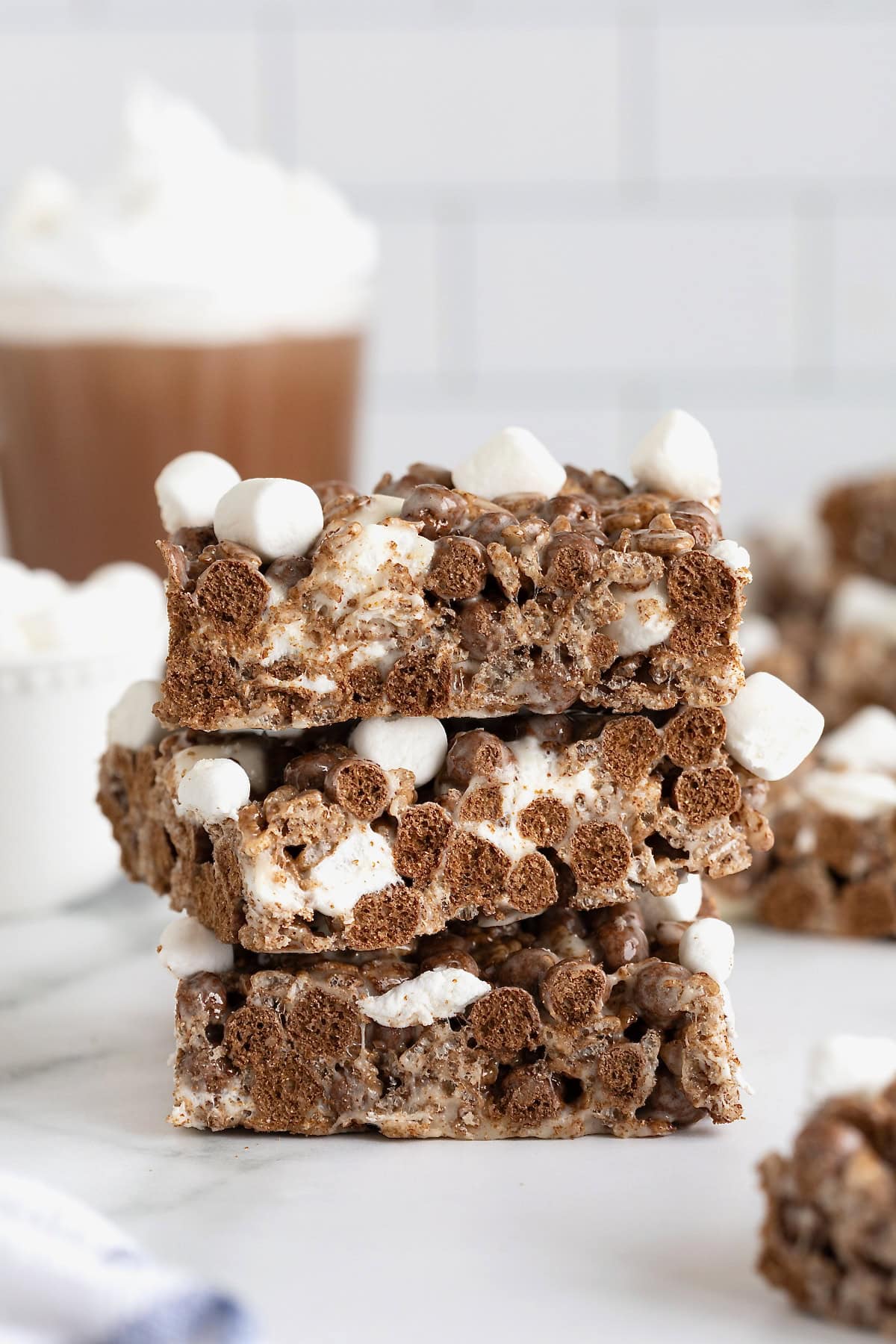 Three no-bake hot cocoa Krispie treats topped with mini marshmallows stacked on a. white surface. A glass mug of hot cocoa sits in the background.