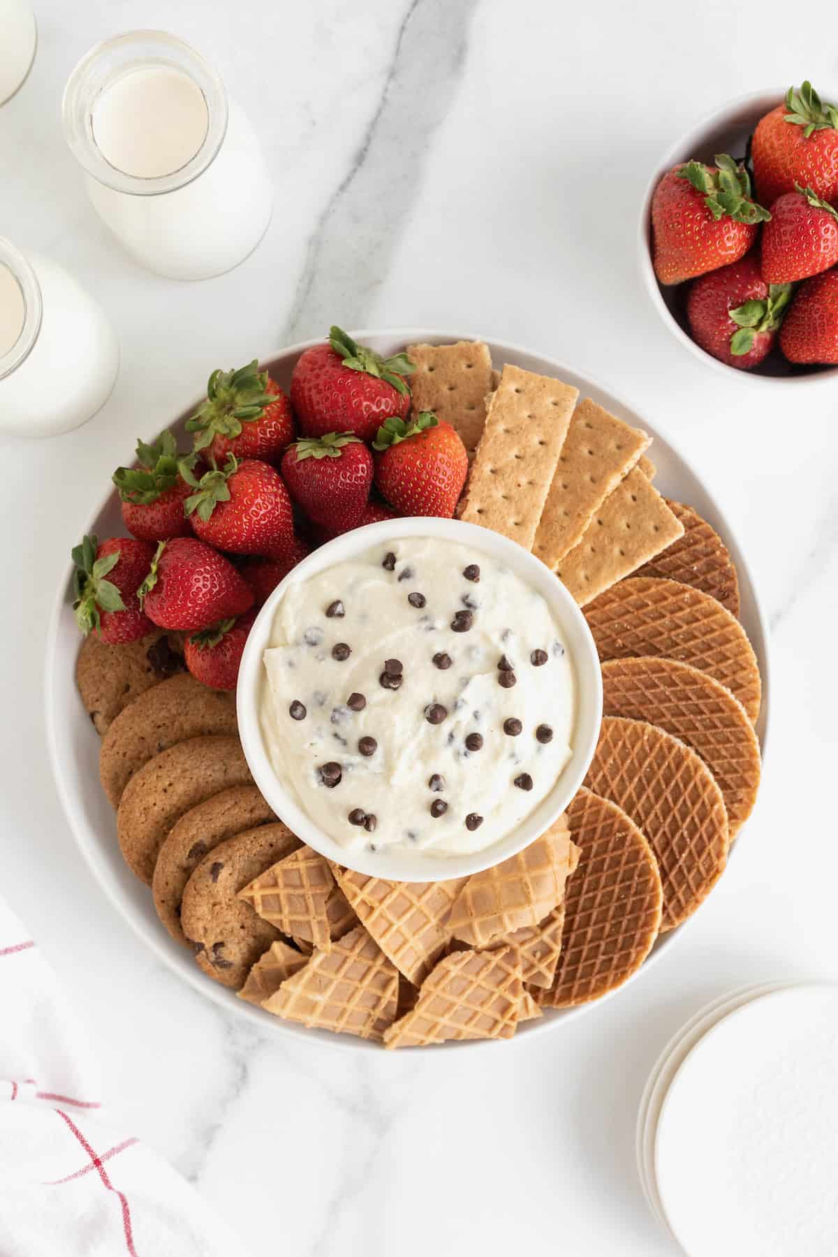 A white serving platter filled with cookies, graham crackers, strawberries and sugar cone wafers. In the center of the dish is a white bowl filled with chocolate chip cannoli dip.