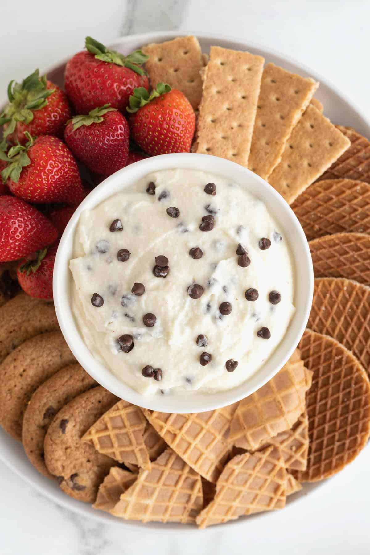 A white serving platter filled with cookies, graham crackers, strawberries and sugar cone wafers. In the center of the dish is a white bowl filled with chocolate chip cannoli dip.