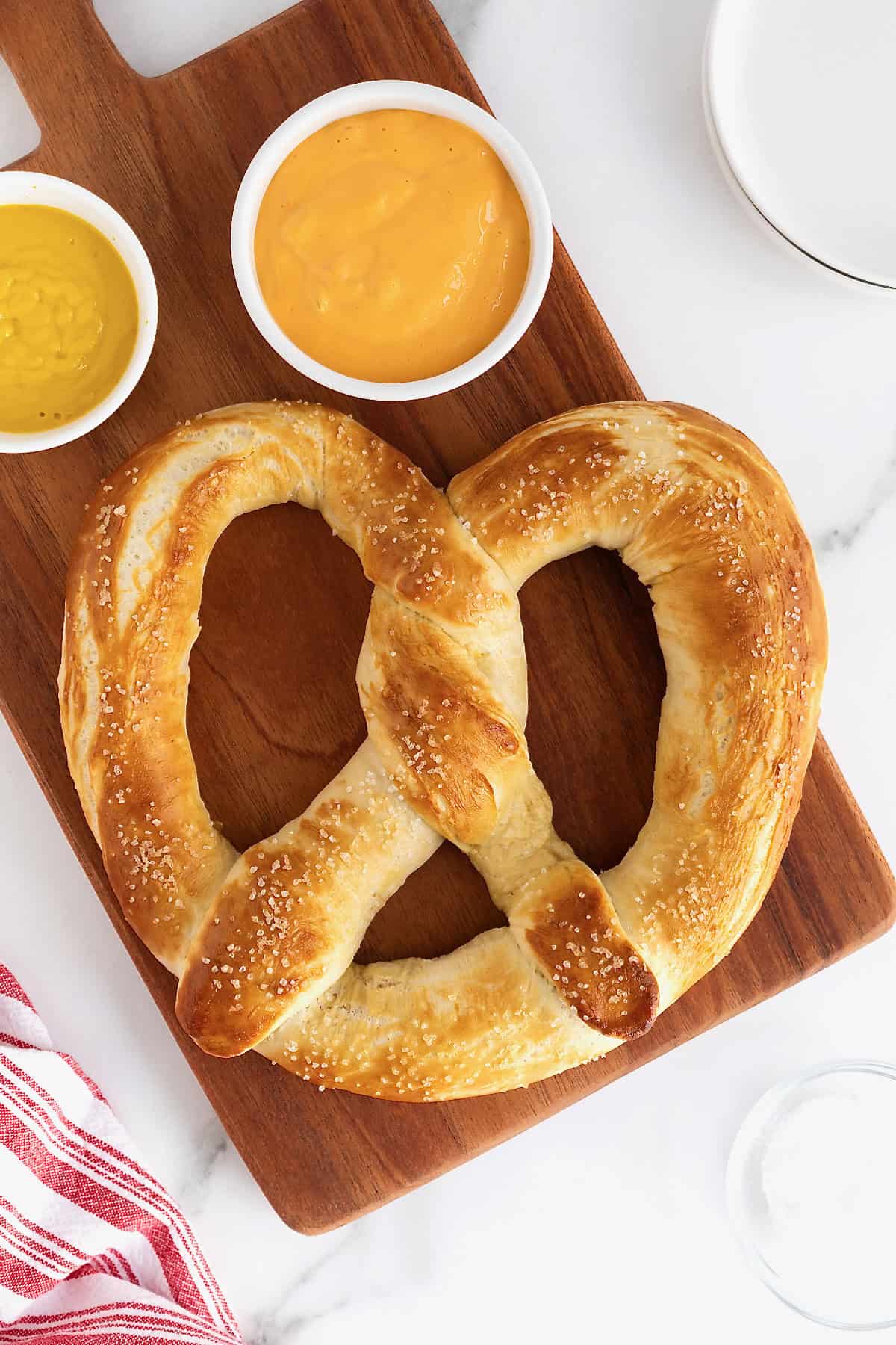 A jumbo pretzel on a walnut wood cutting board next to two small white dishes of mustard and cheese sauces.