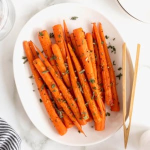 An oblong white serving platter of roasted carrots. A pair of wooden tongs rests on the plate.