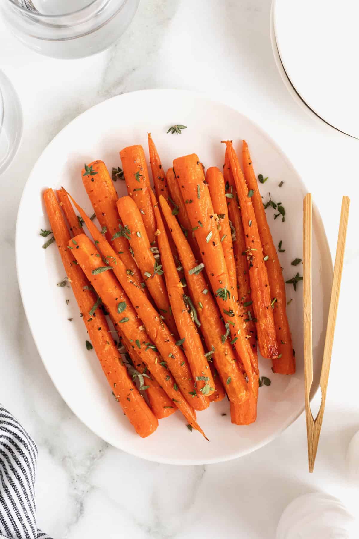 An oblong white serving platter of roasted carrots. A pair of wooden tongs rests on the plate.