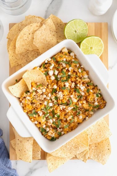 A square white casserole dish of elotes dip surrounded by corn chips on a wooden cutting board.