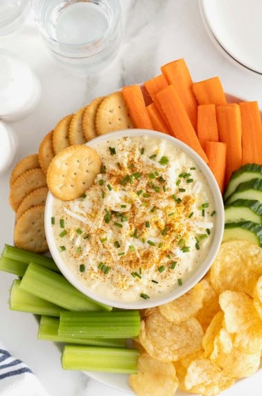 A large white bowl of deviled egg dip surrounded by crackers, carrot sticks, cucumber slices, potato chips and celery sticks on a large round platter.