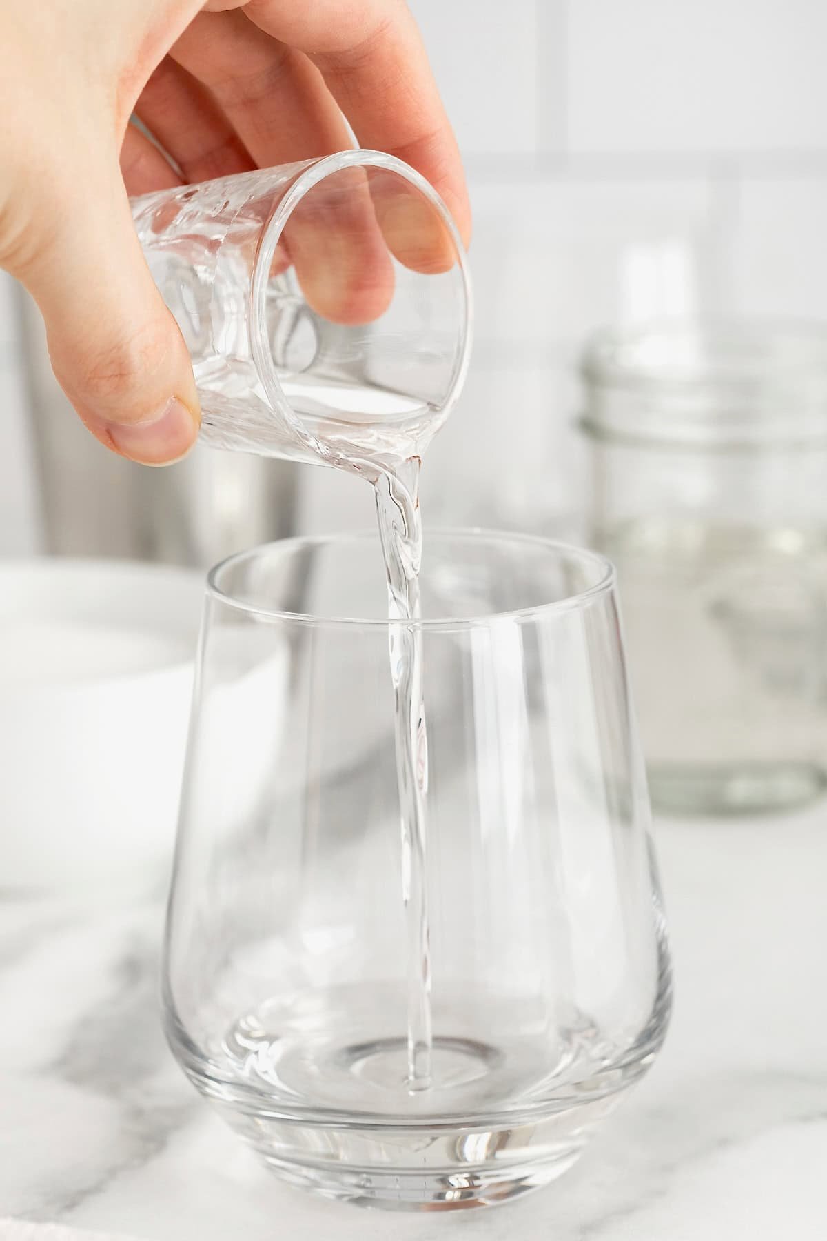 Simple syrup being poured from a glass shot into a glass on a white marble counter.