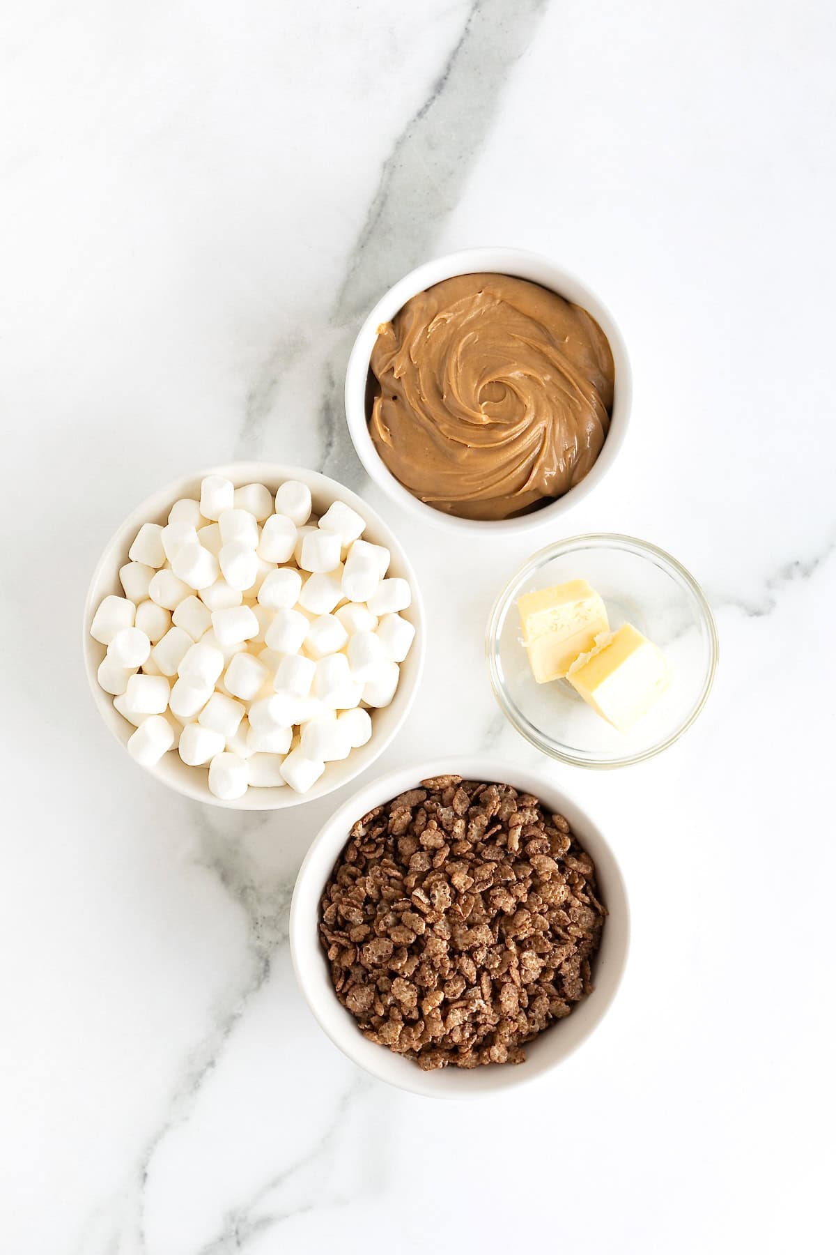 Ingredients to make chocolate peanut butter Rice Krispies treats in small glass dishes on a white marble counter.