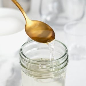 A gold toned spoon pouring simple syrup into a wide mouth glass jar on a white marble counter.