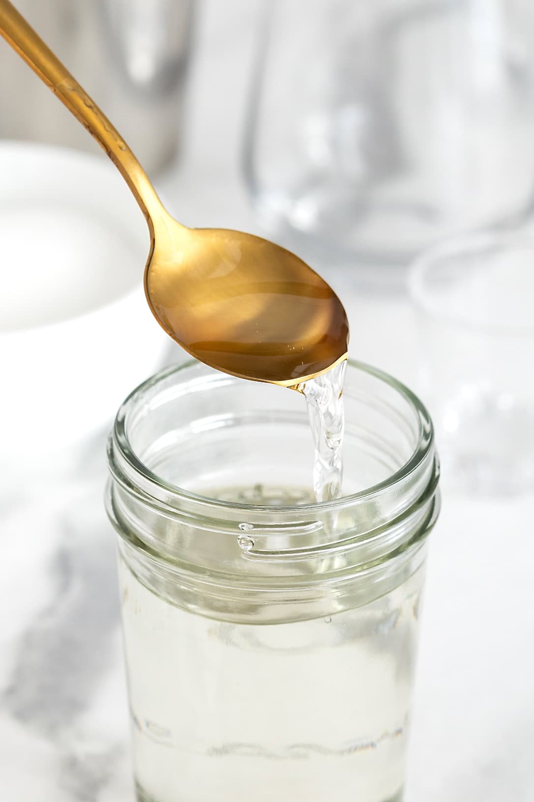 A gold toned spoon pouring simple syrup into a wide mouth glass jar on a white marble counter.