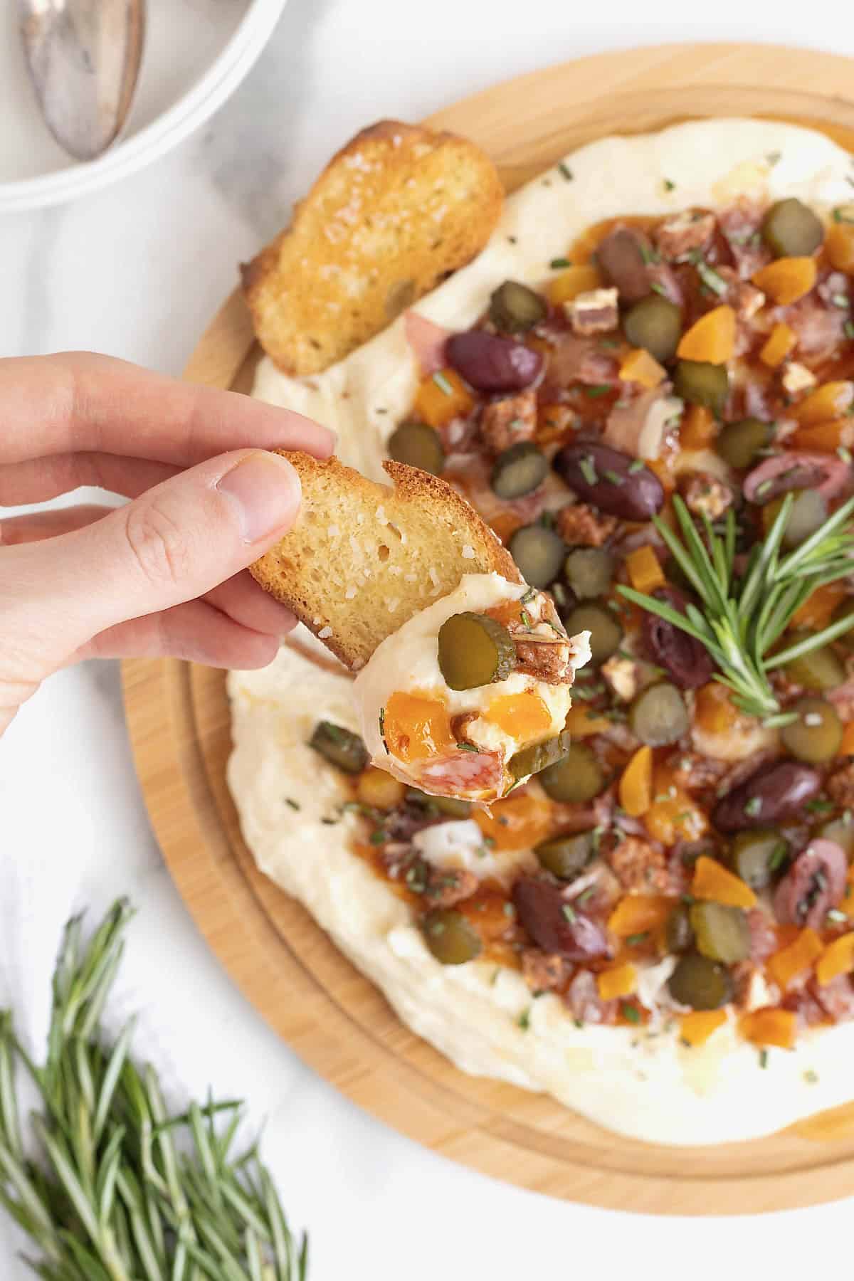 A piece of crostini being dipped into charcuterie dip on a round wooden food board.