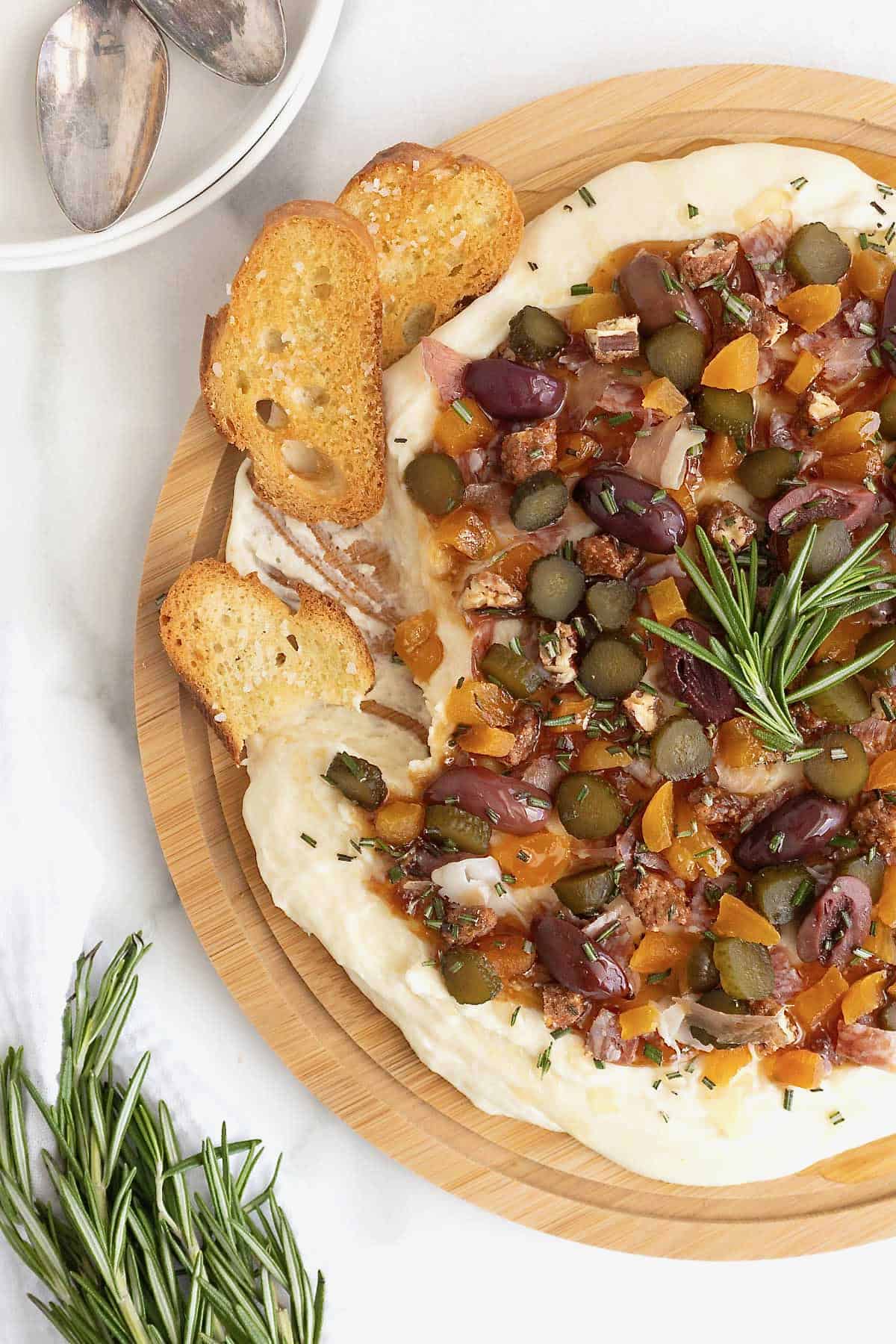 A round wood food board spread with charcuterie dip. Three slices of crostini sit on the top left edge of the food board.