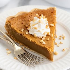 A piece of peanut butter pie topped with a dollop of whipped cream and chopped peanuts on a white plate next to two forks.