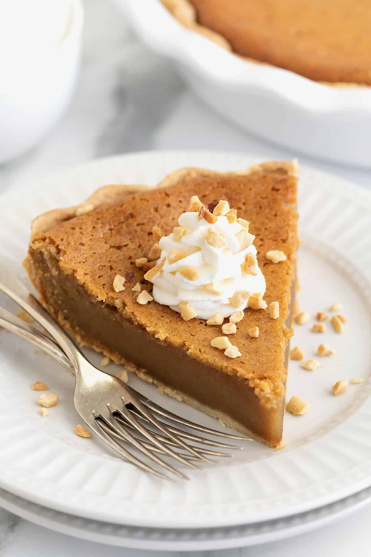 A piece of peanut butter pie topped with a dollop of whipped cream and chopped peanuts on a white plate next to two forks.