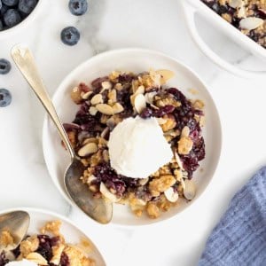 A small white plate of blueberry almond crisp. There is a scoop of vanilla ice-cream on the crisp and a spoon lying next to it.