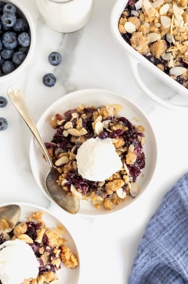 A small white plate of blueberry almond crisp. There is a scoop of vanilla ice-cream on the crisp and a spoon lying next to it.