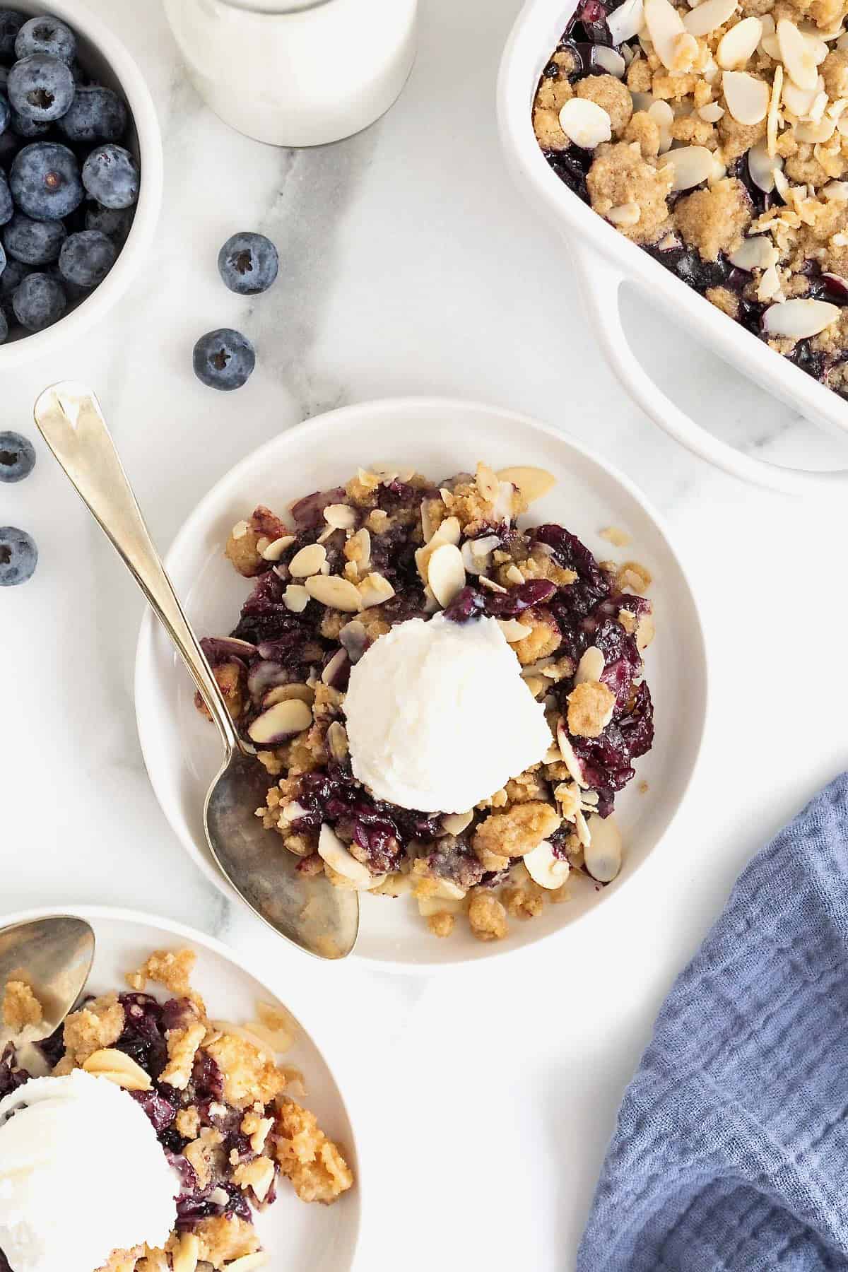 A small white plate of blueberry almond crisp. There is a scoop of vanilla ice-cream on the crisp and a spoon lying next to it.