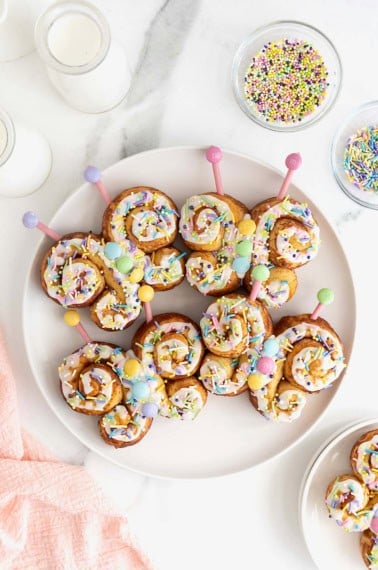 Four butterfly cinnamon rolls decorated with icing and pastel candies on a large white plate.