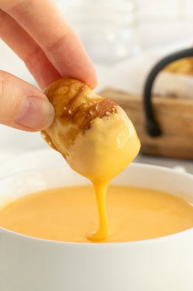 A soft pretzel bite being dipped into a white bowl of cheese sauce.