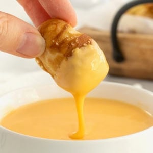 A soft pretzel bite being dipped into a white bowl of cheese sauce.