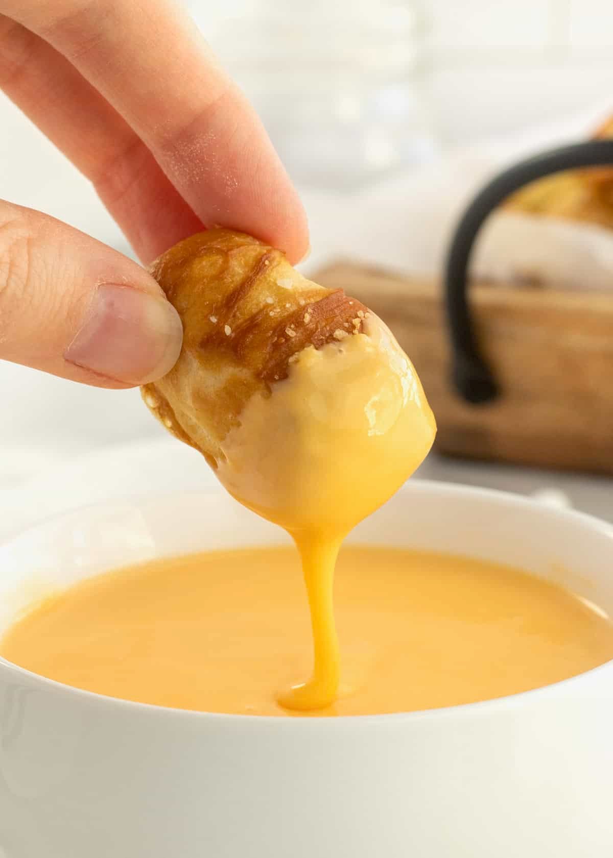 A soft pretzel bite being dipped into a white bowl of cheese sauce.