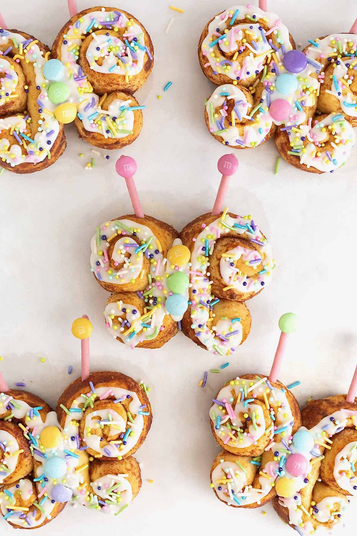Five butterfly shaped cinnamon rolls decorated with icing, pastel candies and candy antennae on a whit surface.