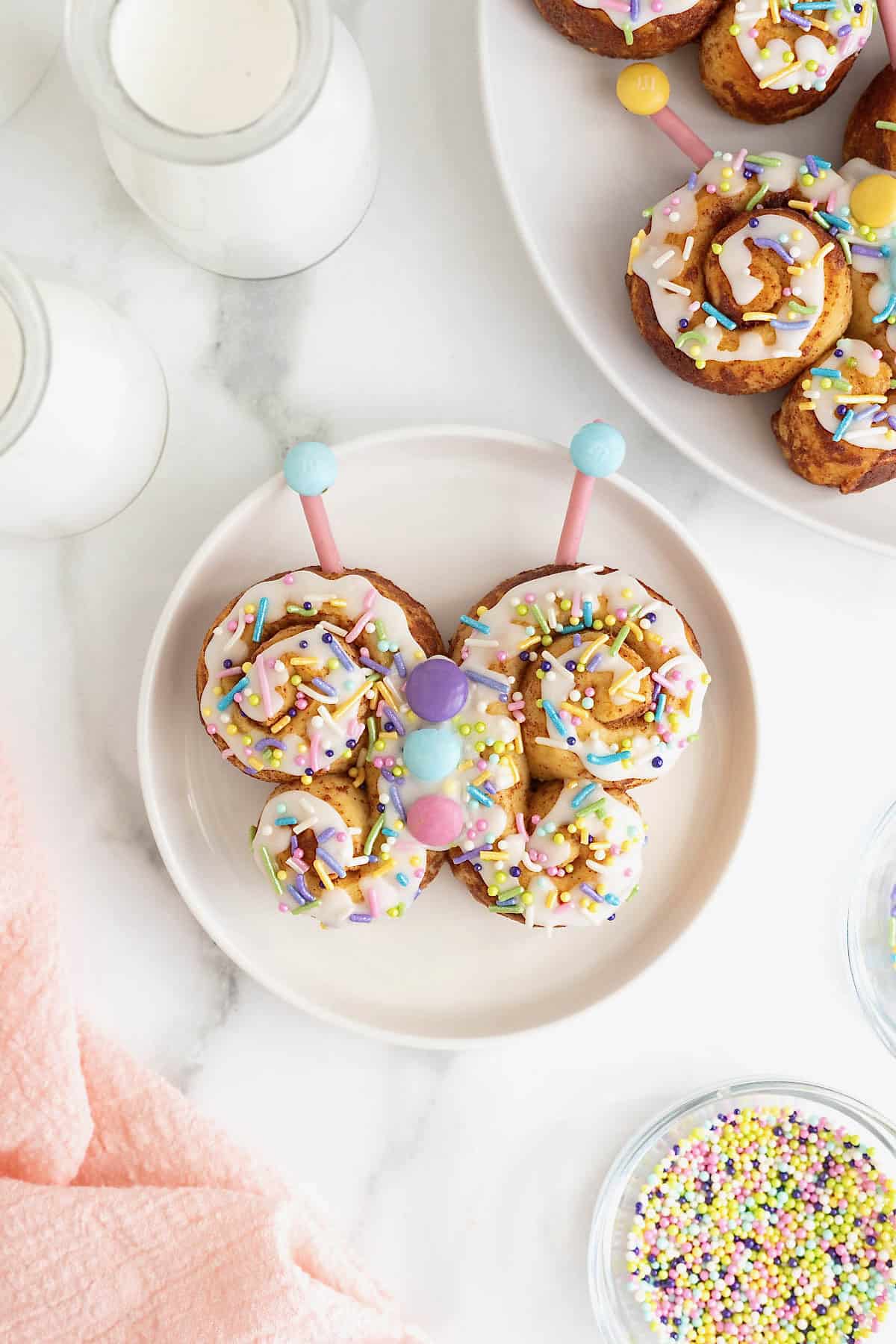 A butterfly-shaped cinnamon roll decorated with white icing and pastel M&Ms and sprinkles on a white plate.