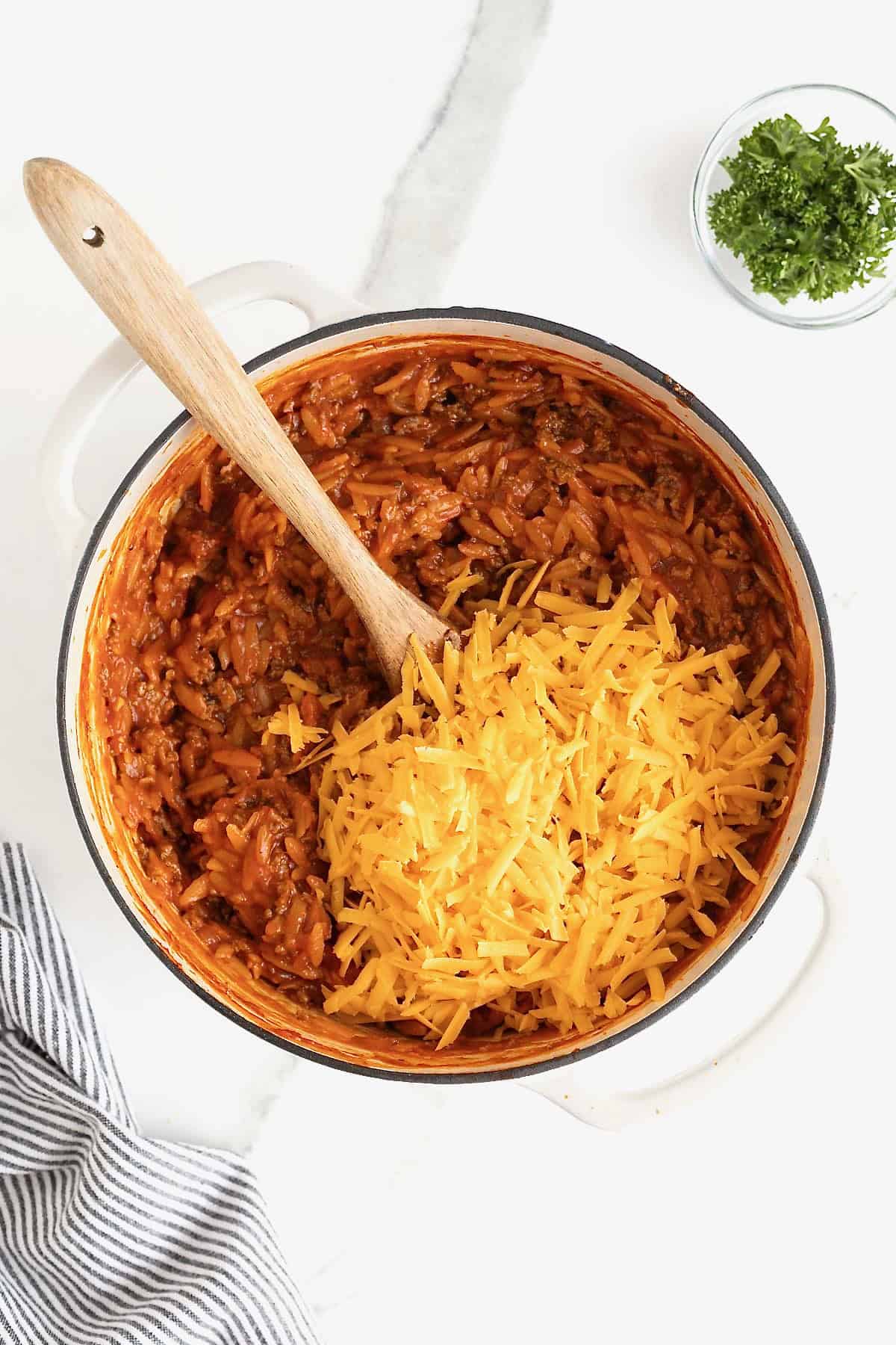 Shredded cheddar cheese added to a white dutch oven of sloppy Joe orzo.