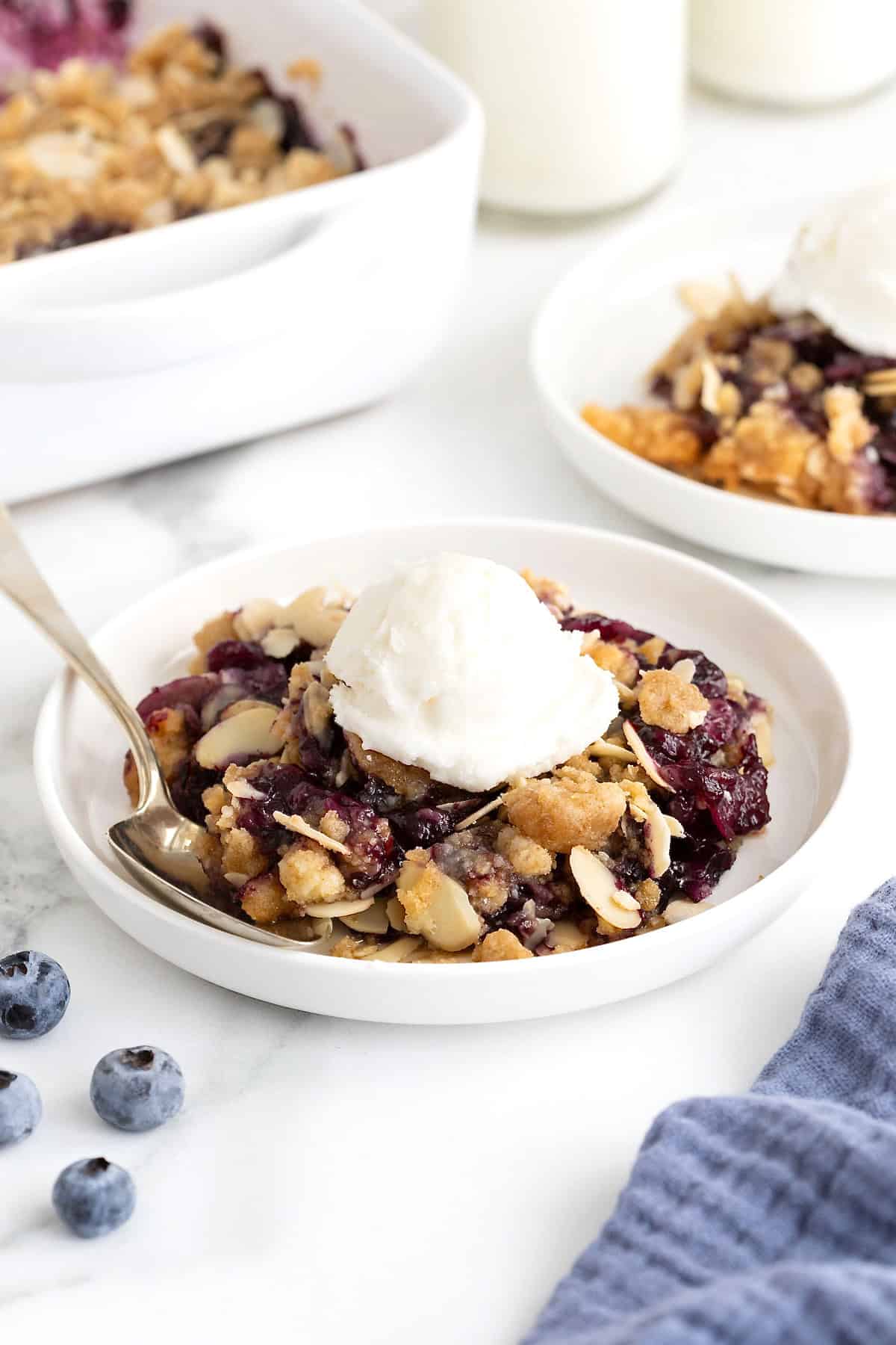 Two small white plates of blueberry almond crisp. There is a scoop of vanilla ice-cream on the crisp and a spoon lying next to it.