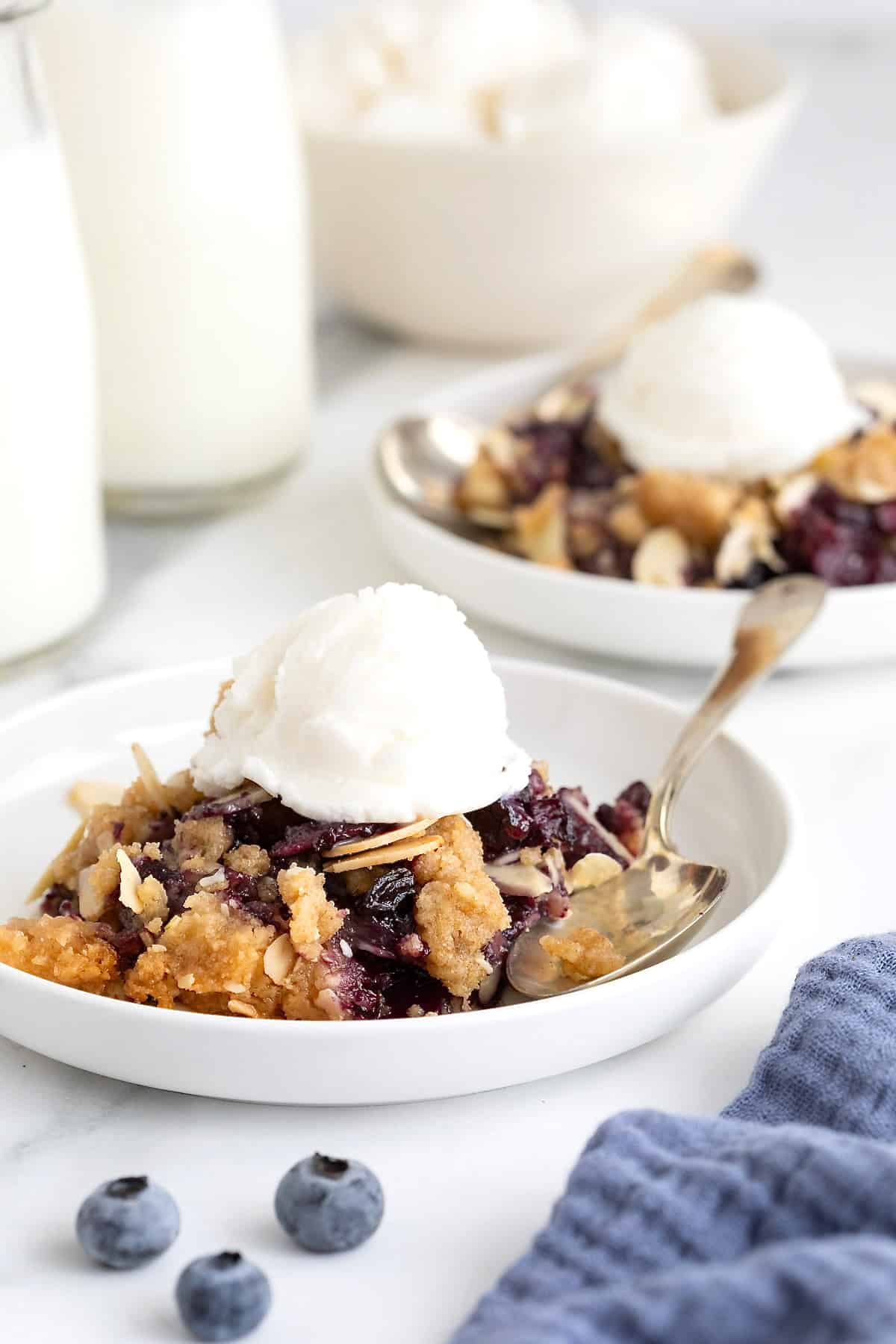 Two small white plates of blueberry almond crisp. There is a scoop of vanilla ice-cream on the crisp and a spoon lying next to it.