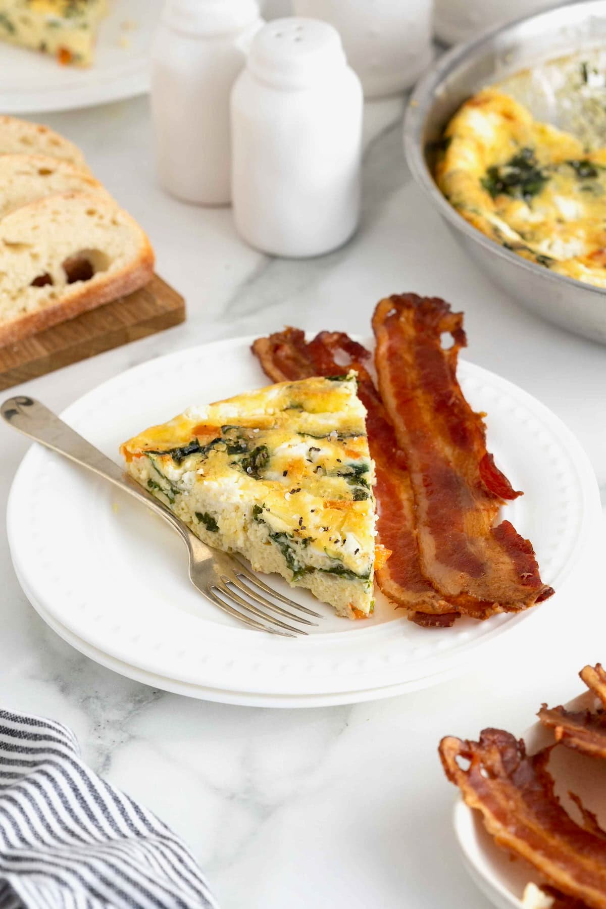 A slice of frittata on a white plate next to two strips of bacon.