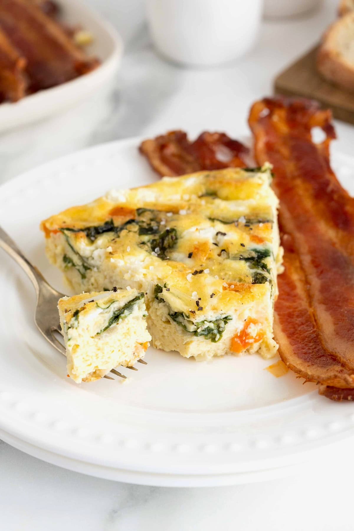 A slice of frittata on a white plate next to three strips of bacon.