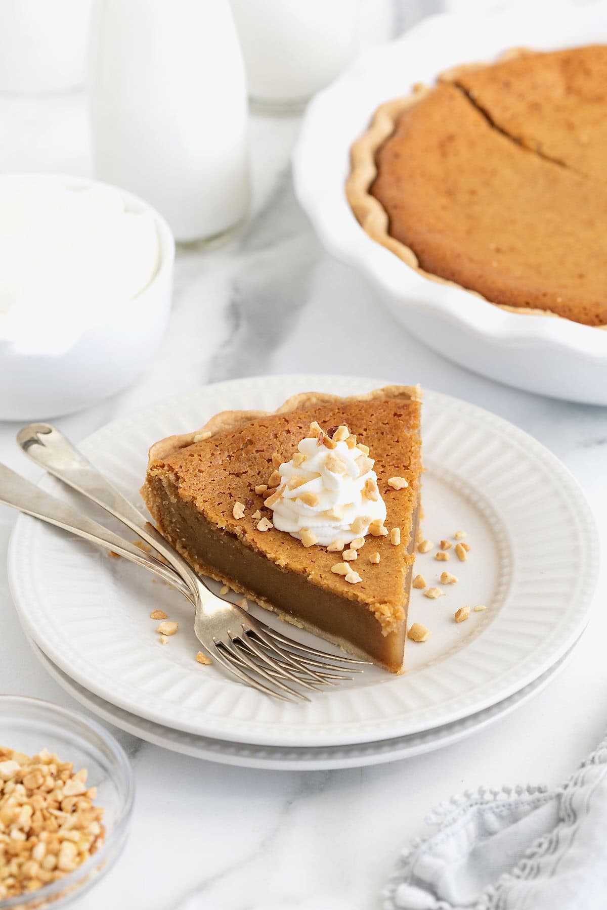 A piece of peanut butter pie topped with a dollop of whipped cream and chopped peanuts on a white plate next to two forks.