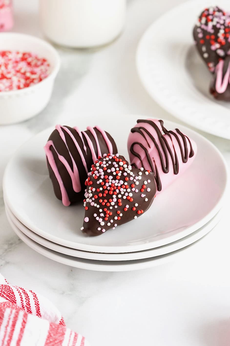 Three heart-shaped chocolate strawberries decorated with Valentine sprinkles on a stack of three white plates.