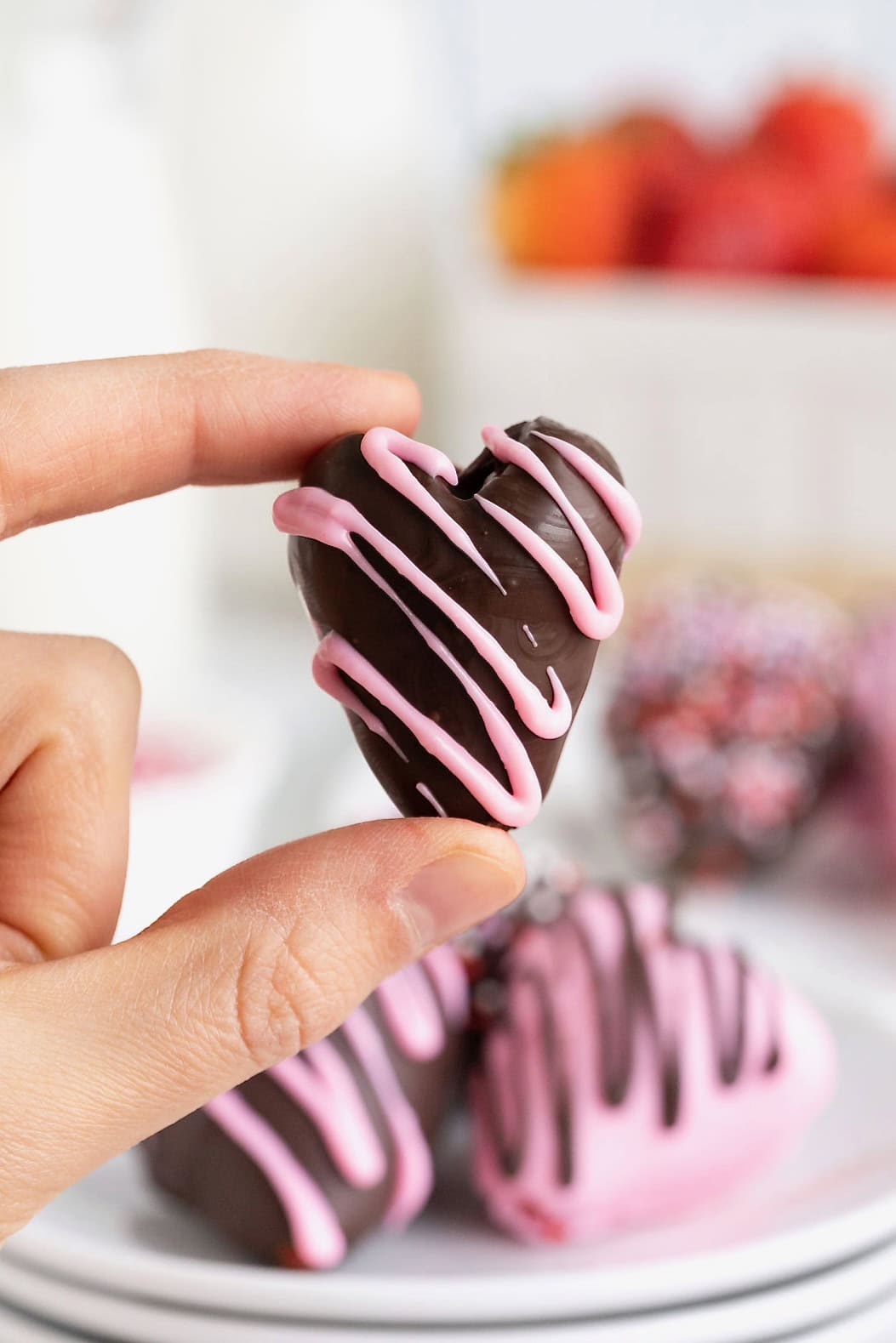 A heart-shaped chocolate covered strawberry held between a thumb and forefinger.