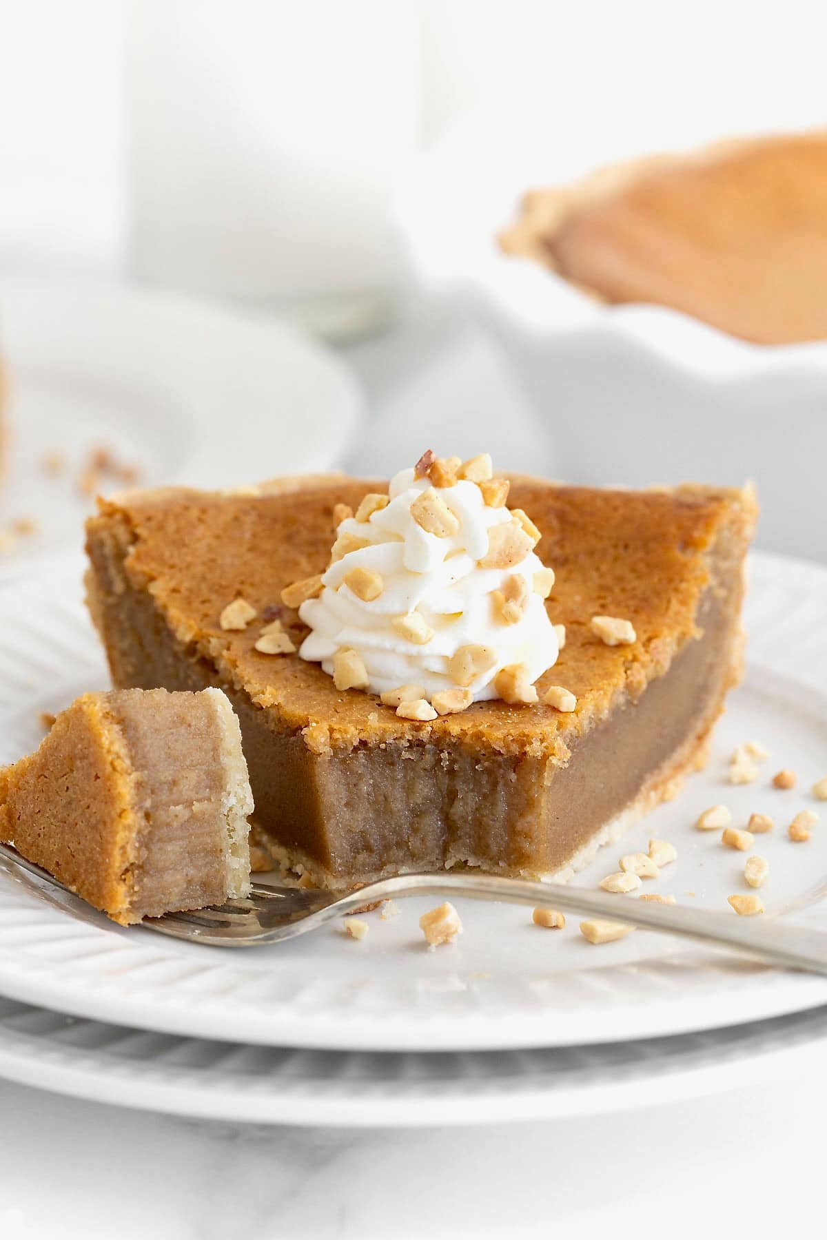 A piece of peanut butter pie topped with a dollop of whipped cream and chopped peanuts on a white plate next to a bite of pie on a fork.