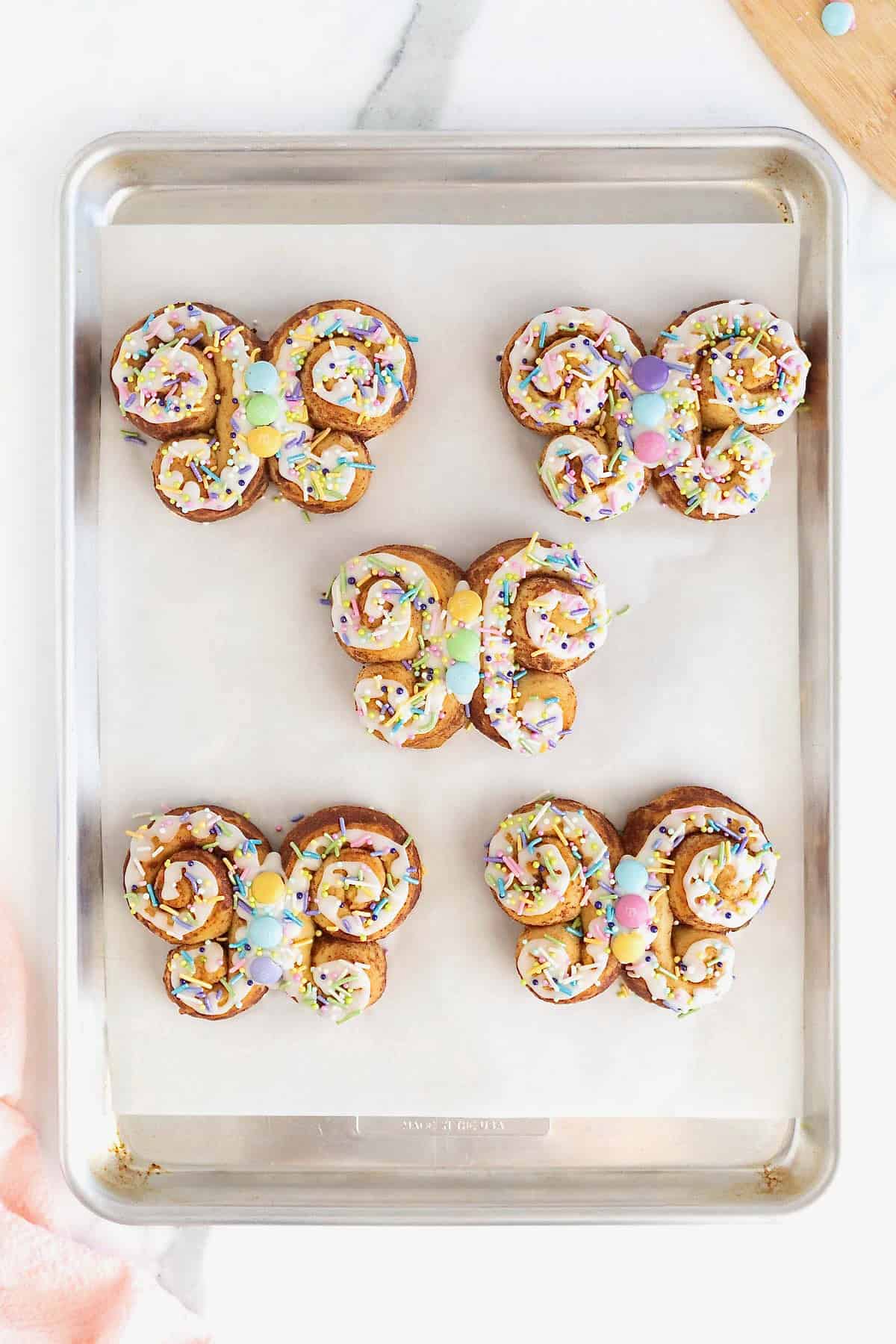 Five butterfly shaped cinnamon rolls decorated with white icing and pastel candies on a parchment lined aluminum baking sheet.