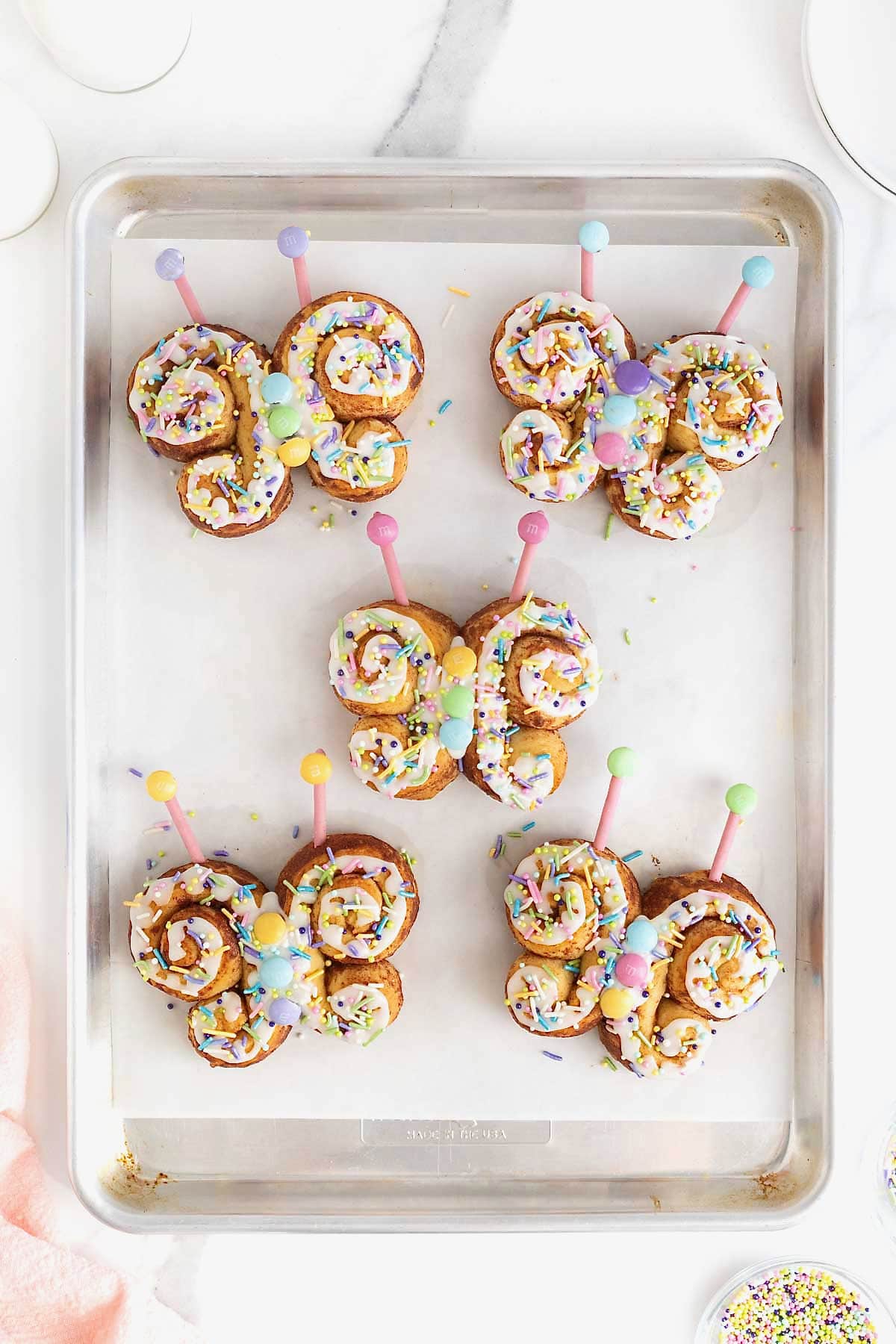Five butterfly shaped cinnamon rolls decorated with white icing and pastel candies on a parchment lined aluminum baking sheet.