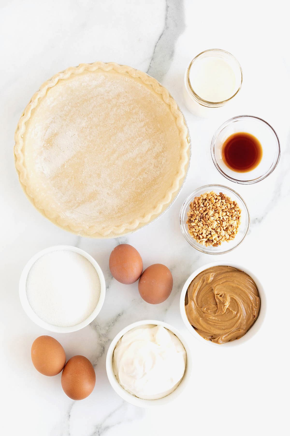 Ingredients for baked peanut butter pie in small glass dishes on a white marble counter next to an unbaked pie crust with fluted edges.