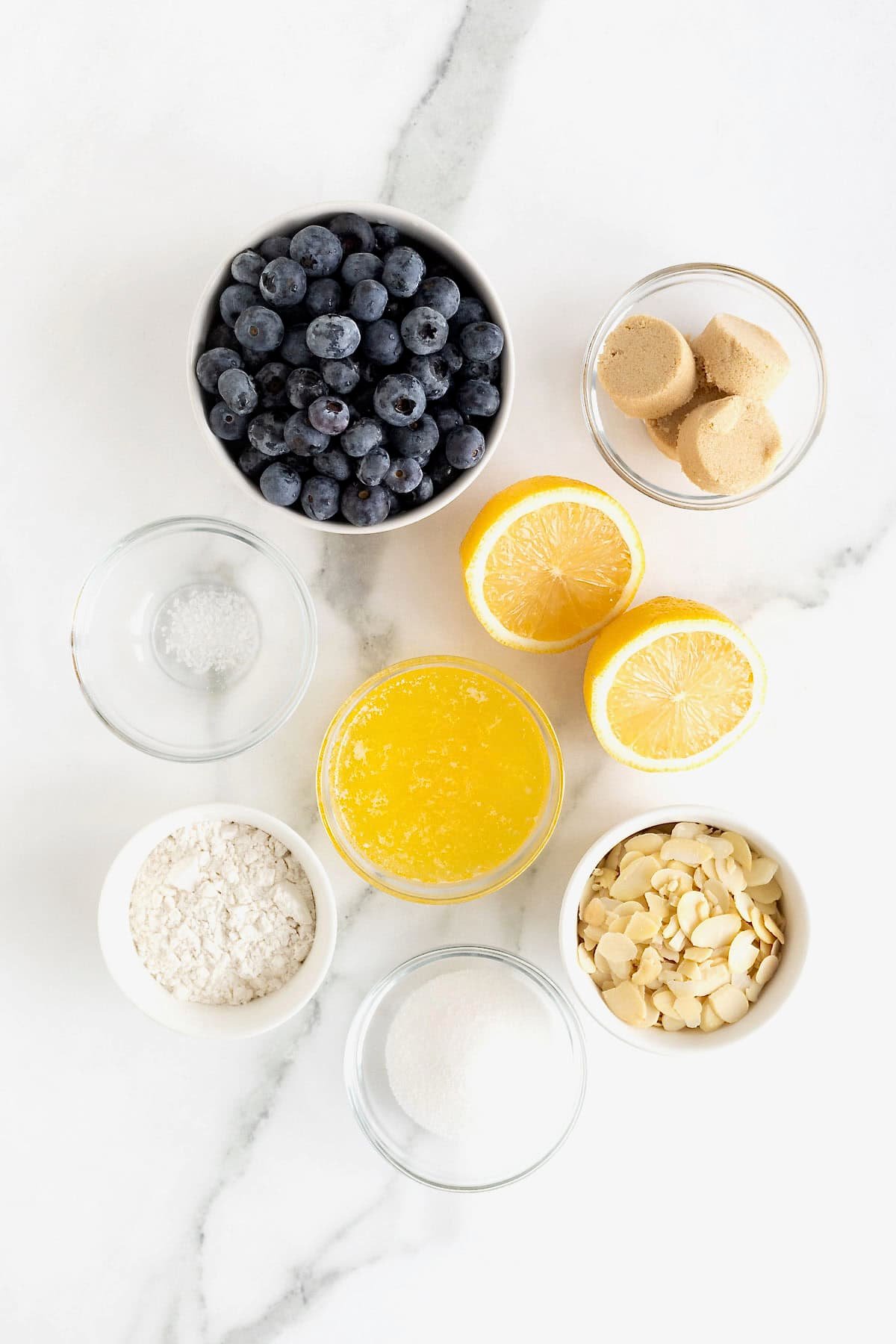 Ingredients to make blueberry almond crisp in small glass dishes on a white marble counter.