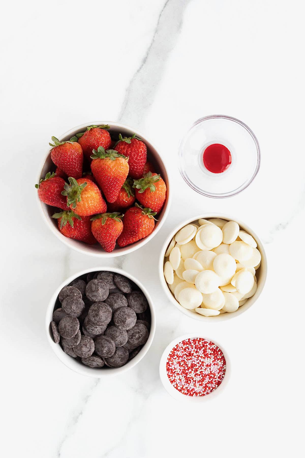 Ingredients to make heart-shaped chocolate covered strawberries in small glass dishes on a white marble counter.