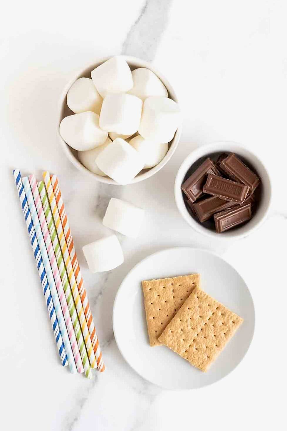 Ingredients to make s'mores marshmallow pops in white dishes on a white marble counter.