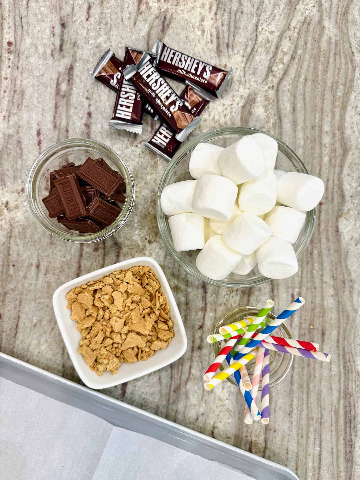 Ingredients for S'mores Marshmallow Pops in small dishes on a dark grey marble counter.