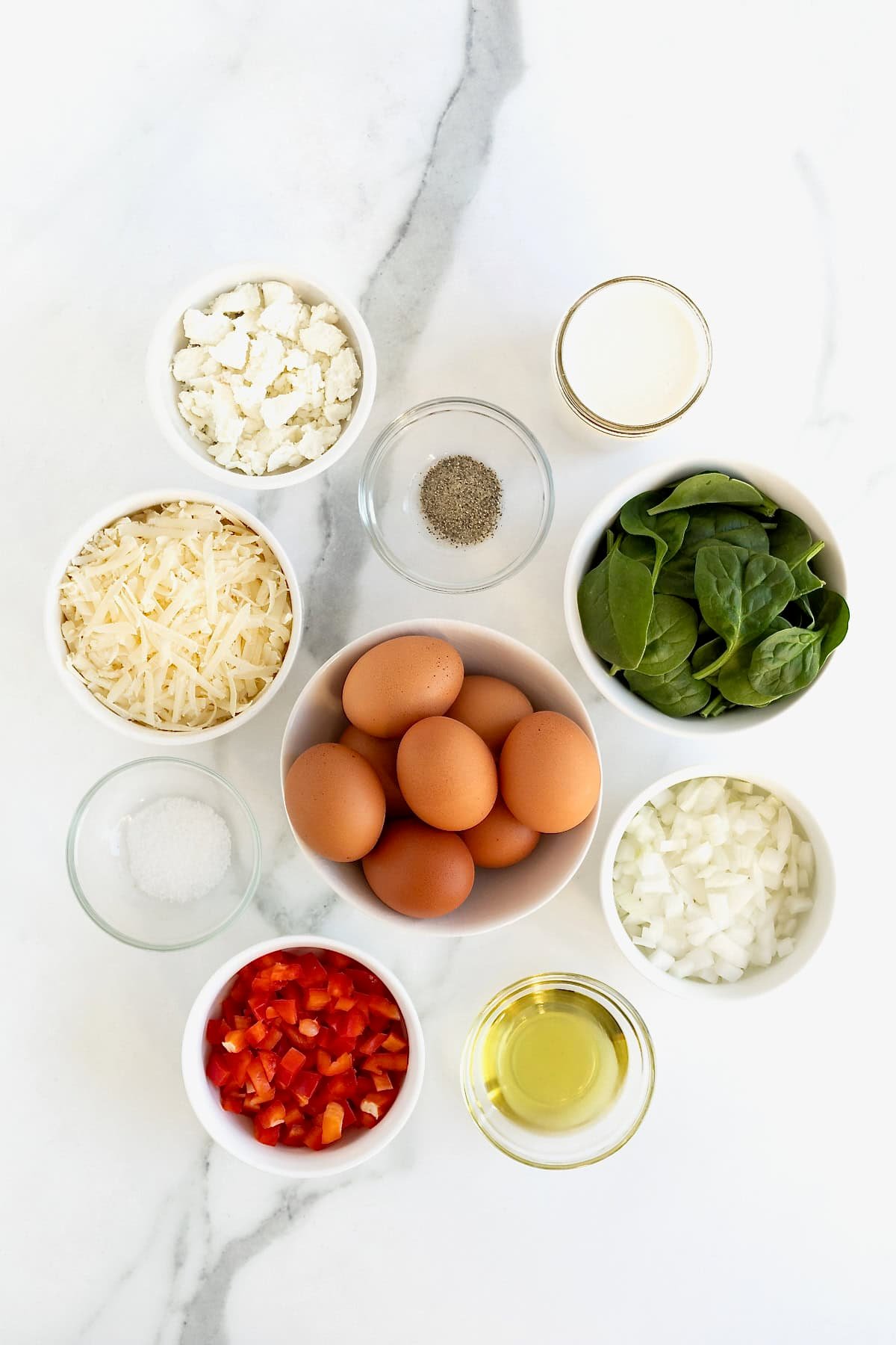 Ingredients to make spinach, red pepper and goat cheese frittata in small glass dish on a white marble counter.