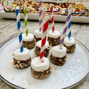 Eight large marshmallows with straws sticking out on a large plate. The marshmallows have been dipped halfway in melted chocolate and rolled in graham cracker crumbs.