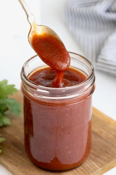 A spoon being dipped into a wide mouth mason jar of barbecue sauce on a wooden cutting board.
