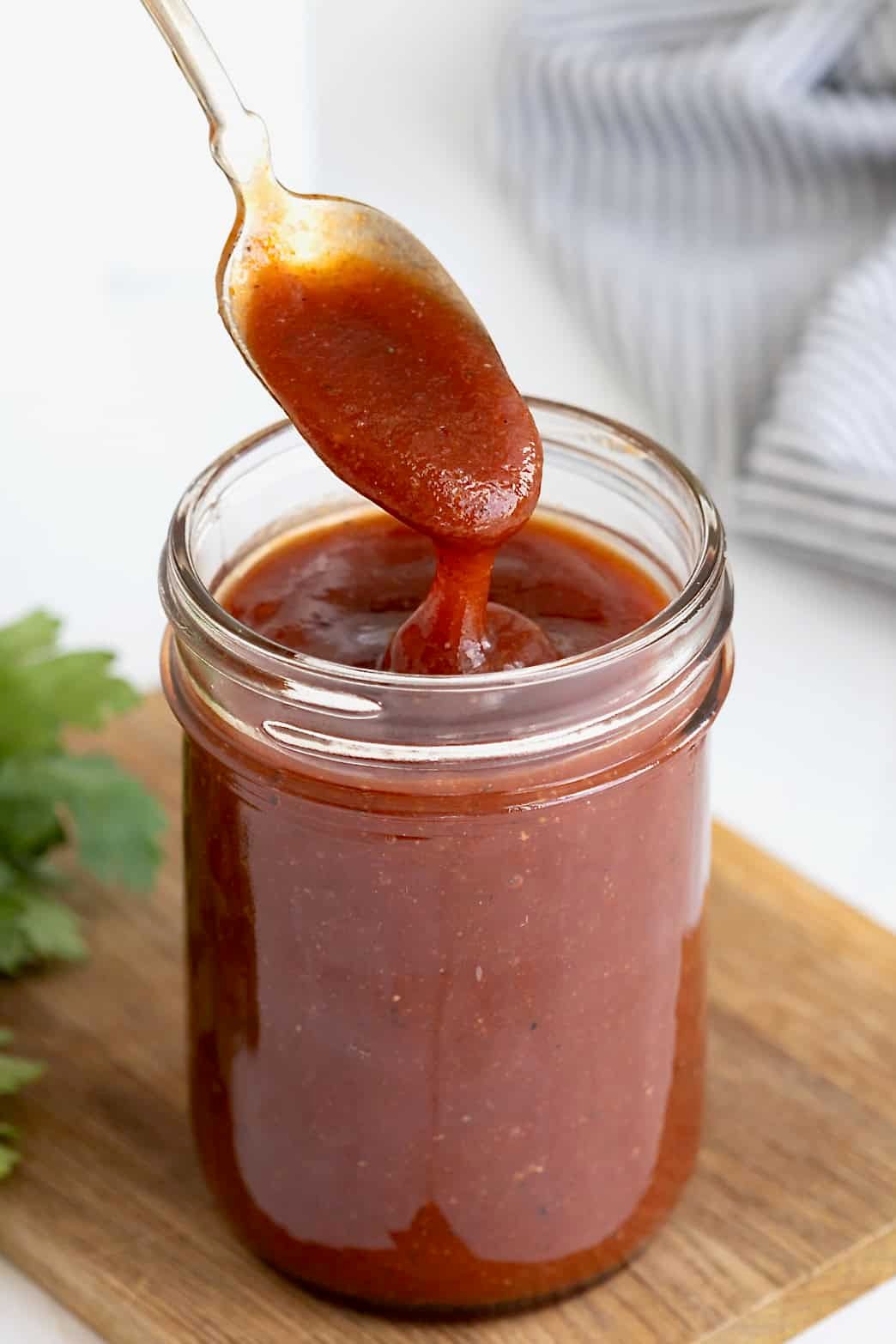 A spoon being dipped into a wide mouth mason jar of barbecue sauce on a wooden cutting board.