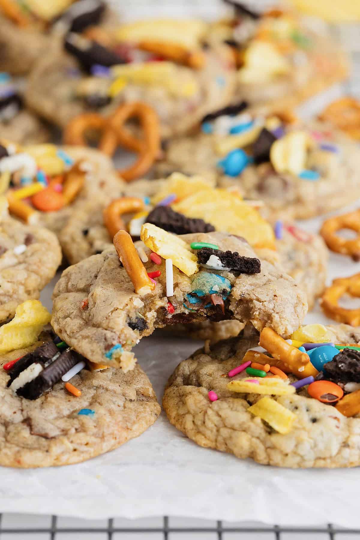 A pile of cookies with pretzel, potato chip, Oreo bits and M&Ms in them. There is a bite out of the top cookie.
